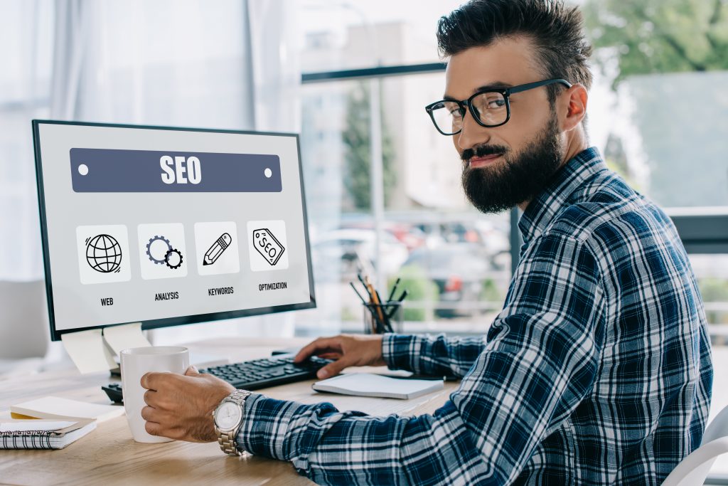 young successful seo manager sitting at workplace with computer and looking at camera